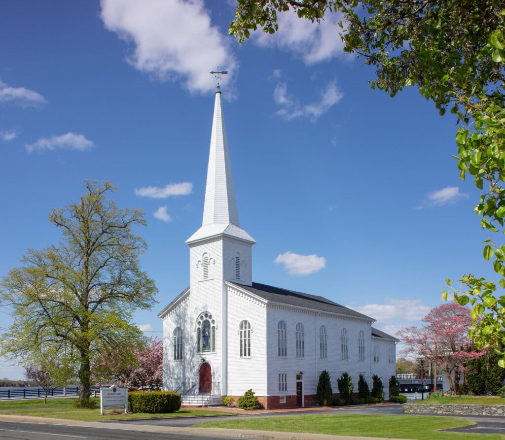 Beautiful white wooden church with a tall steeple under a blue sky, surrounded by blooming trees and lush green grass, showcasing expert construction and roofing services.