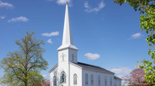 Beautiful white wooden church with a tall steeple under a blue sky, surrounded by blooming trees and lush green grass, showcasing expert construction and roofing services.